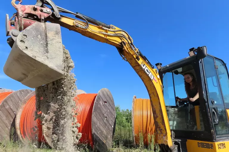 Baggerfahrerin bei Tiefbauarbeiten für Glasfaser, daneben Trommeln mit orangen Leerrohren